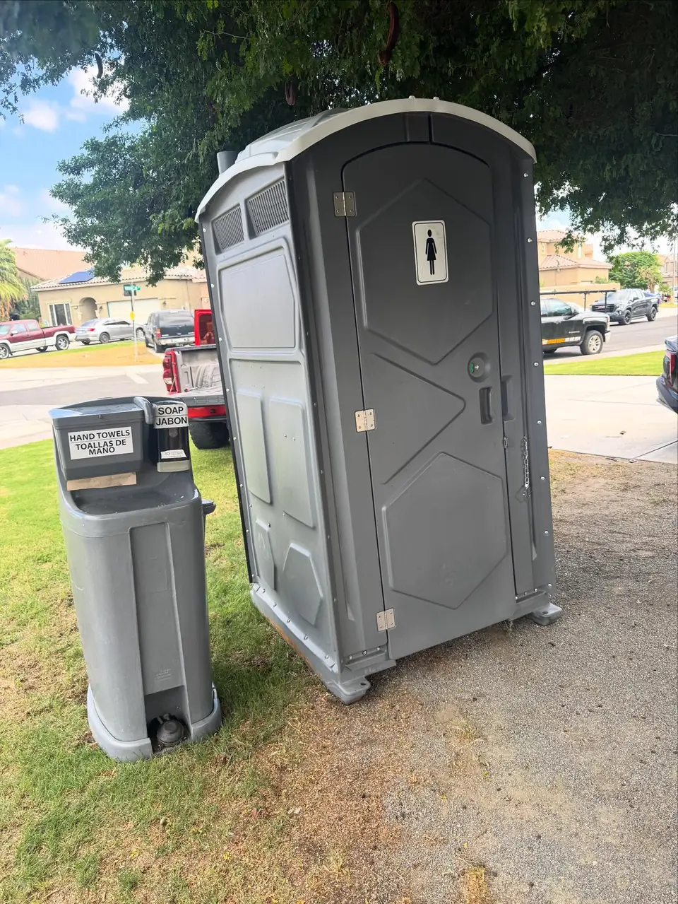 Gray standard porta potty beside a foot-pump hand wash station on a grass lawn